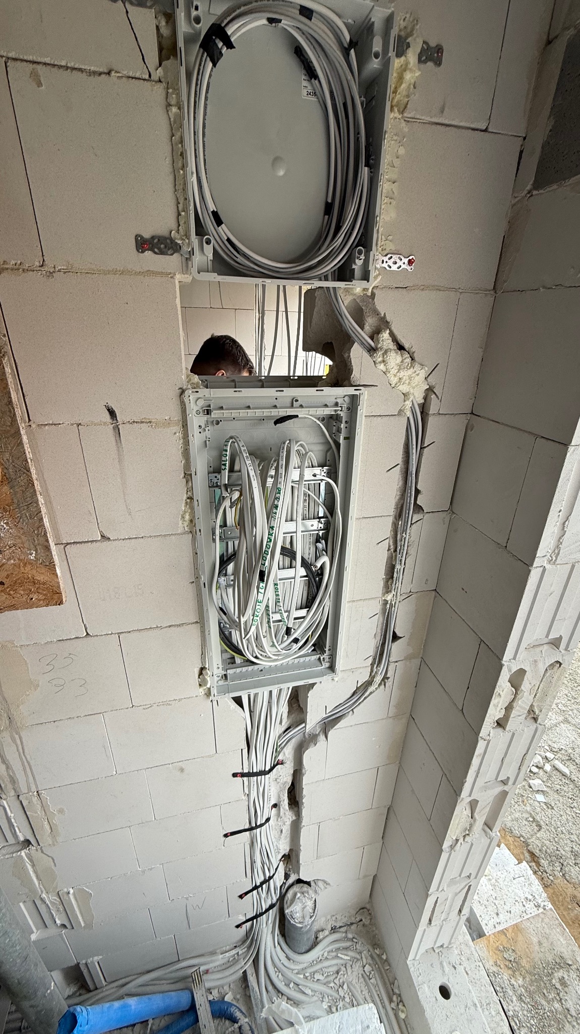 Rozdzielnia elektryczna w trakcie montażu w ścianie z betonu komórkowego. Widoczne liczne przewody i fragment głowy elektryka w otworze.