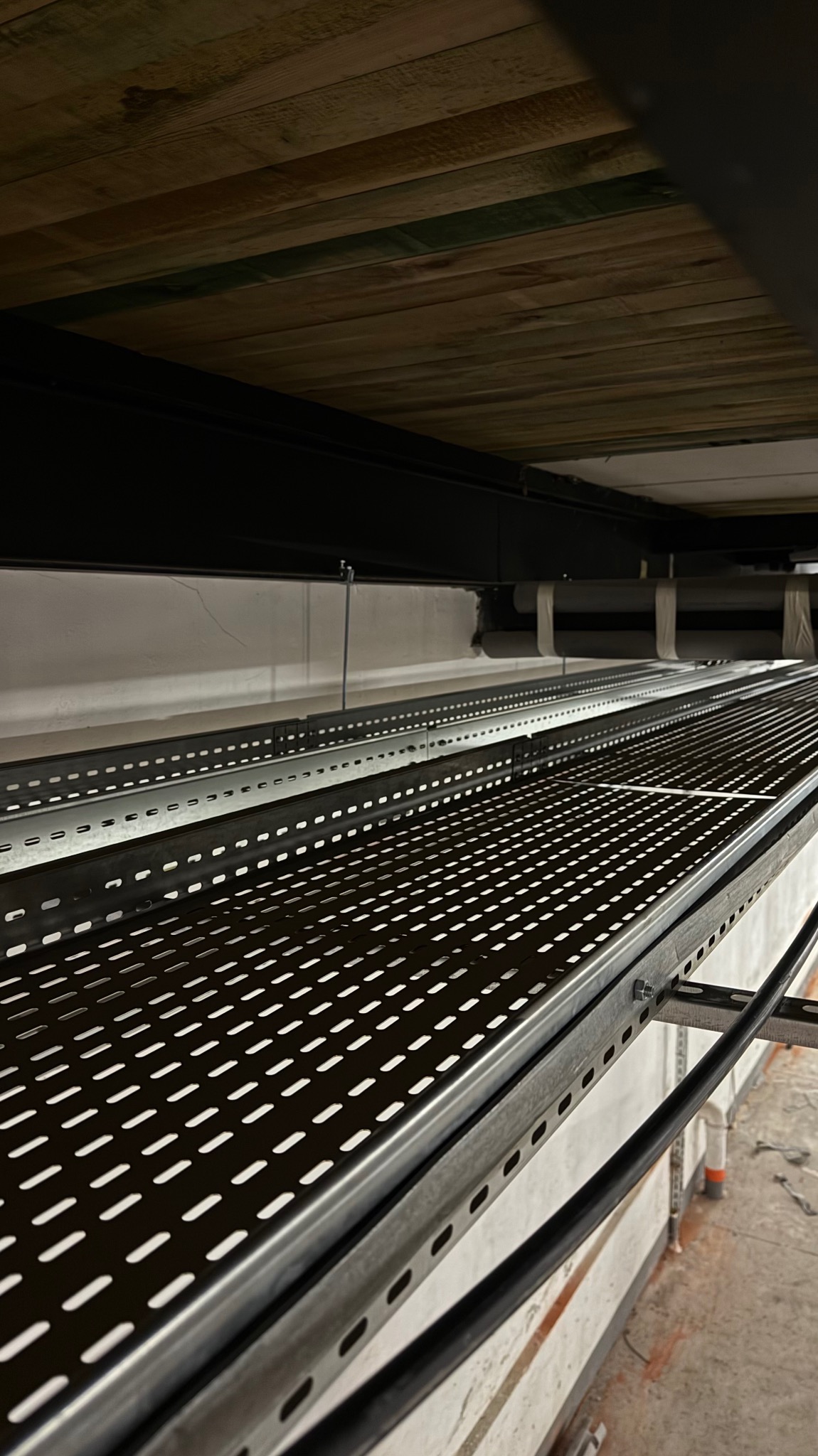 Metalowe korytka kablowe zamontowane pod sufitem z drewnianych desek, widoczne kable. Perspektywa z dołu, oświetlenie sufitowe.