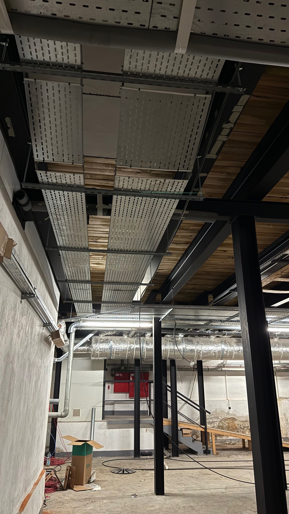 Sufit z instalacjami: koryta kablowe z perforowanej blachy, rury wentylacyjne w izolacji, konstrukcja stalowa. Widok z dołu na sufit w trakcie prac instalacyjnych.