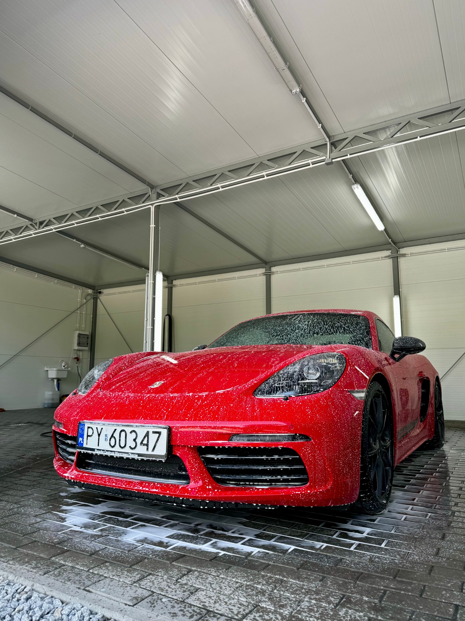 Czerwone Porsche 718 Cayman w trakcie mycia, pokryte pianą, widoczne tablice rejestracyjne PY 60347, myjnia z metalową konstrukcją i oświetleniem LED.