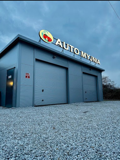 Budynek myjni samochodowej z dwoma bramami wjazdowymi, szarym sidingiem i dużym podświetlanym szyldem z nazwą i logo na tle pochmurnego nieba.