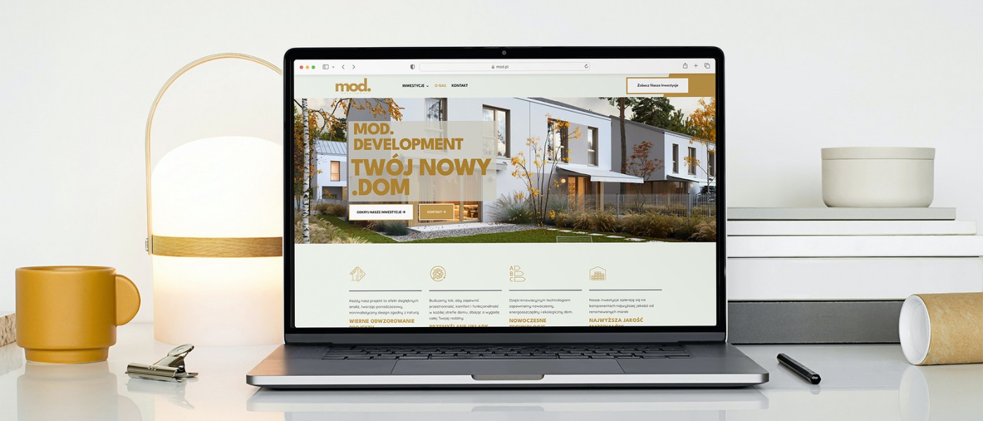 Laptop wyświetla stronę internetową firmy budowlanej z nowoczesnym domem. Minimalistyczny design, stonowana kolorystyka, lampa i kubek w tle.