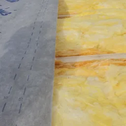 Układanie izolacji z wełny mineralnej i membrany dachowej na drewnianej konstrukcji dachu, widok z góry na częściowo ukończony etap prac.