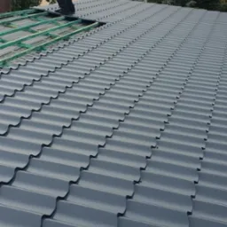 Dekarz montuje szare panele dachowe na konstrukcji drewnianej, widoczny budynek w tle i pochmurne niebo.