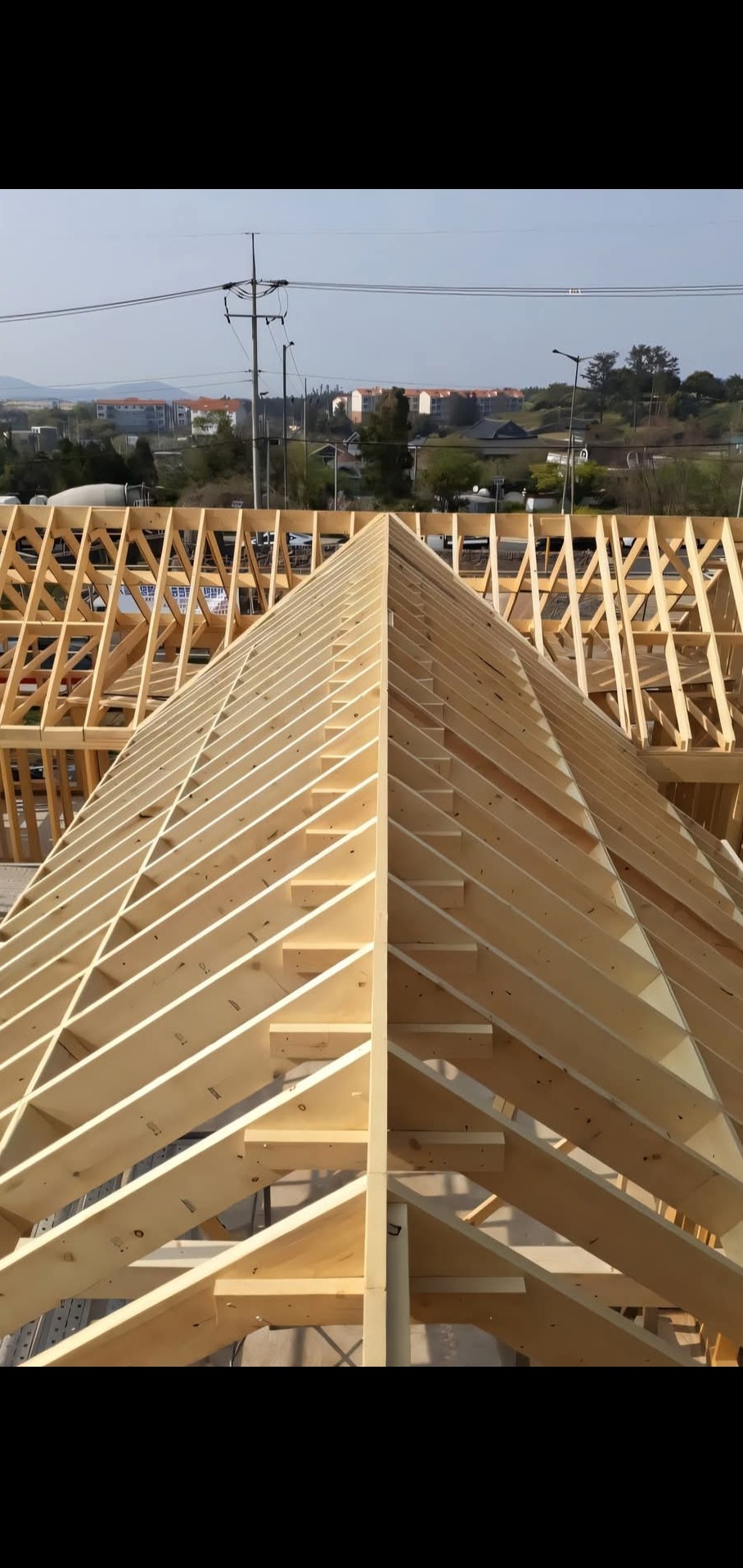 Drewniana konstrukcja dachu w trakcie budowy, widok z góry na szczyt, w tle zabudowa miejska i wzgórza.