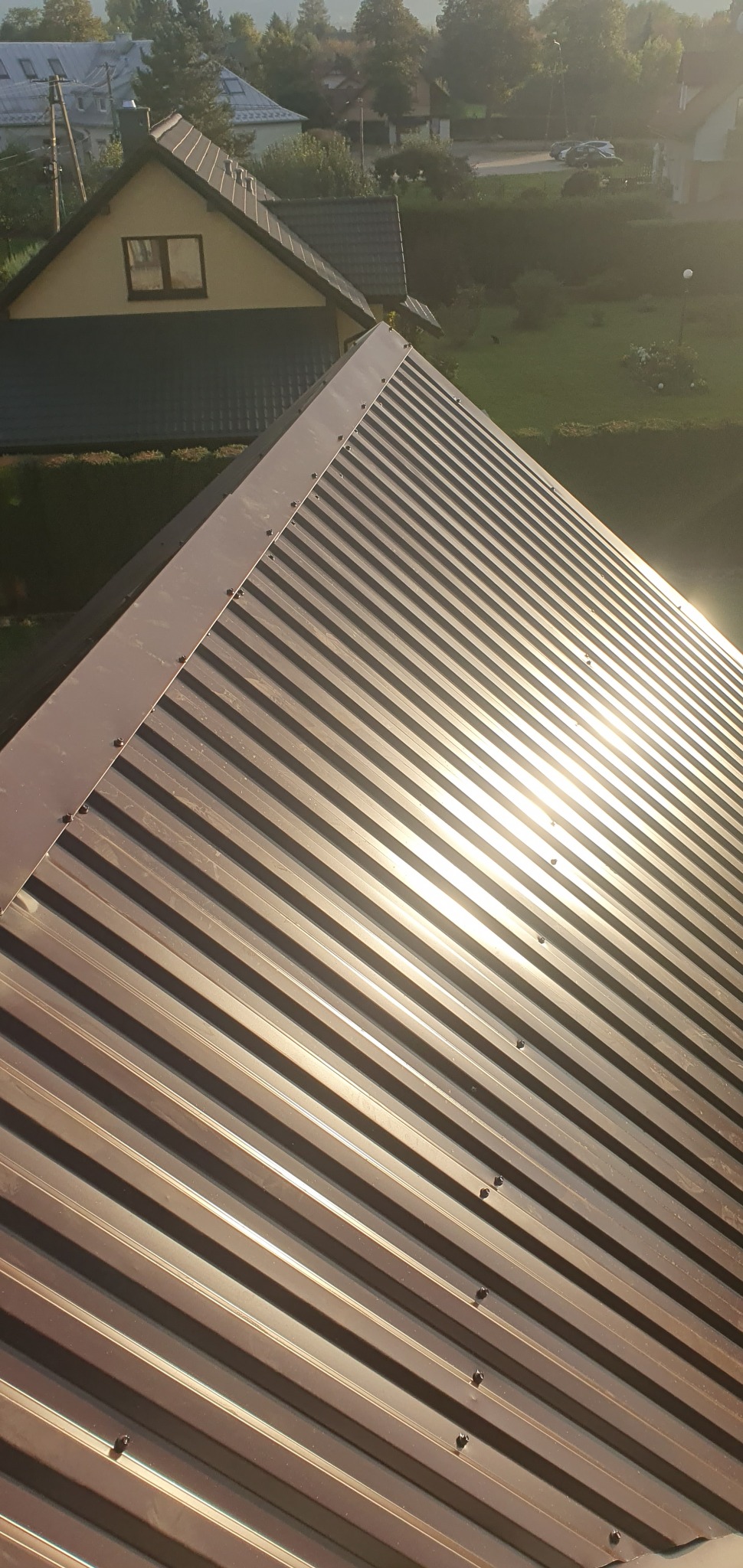 Nowe brązowe krycie dachu z blachy trapezowej, widoczne śruby mocujące, w tle zieleń i zabudowania w słoneczny dzień.