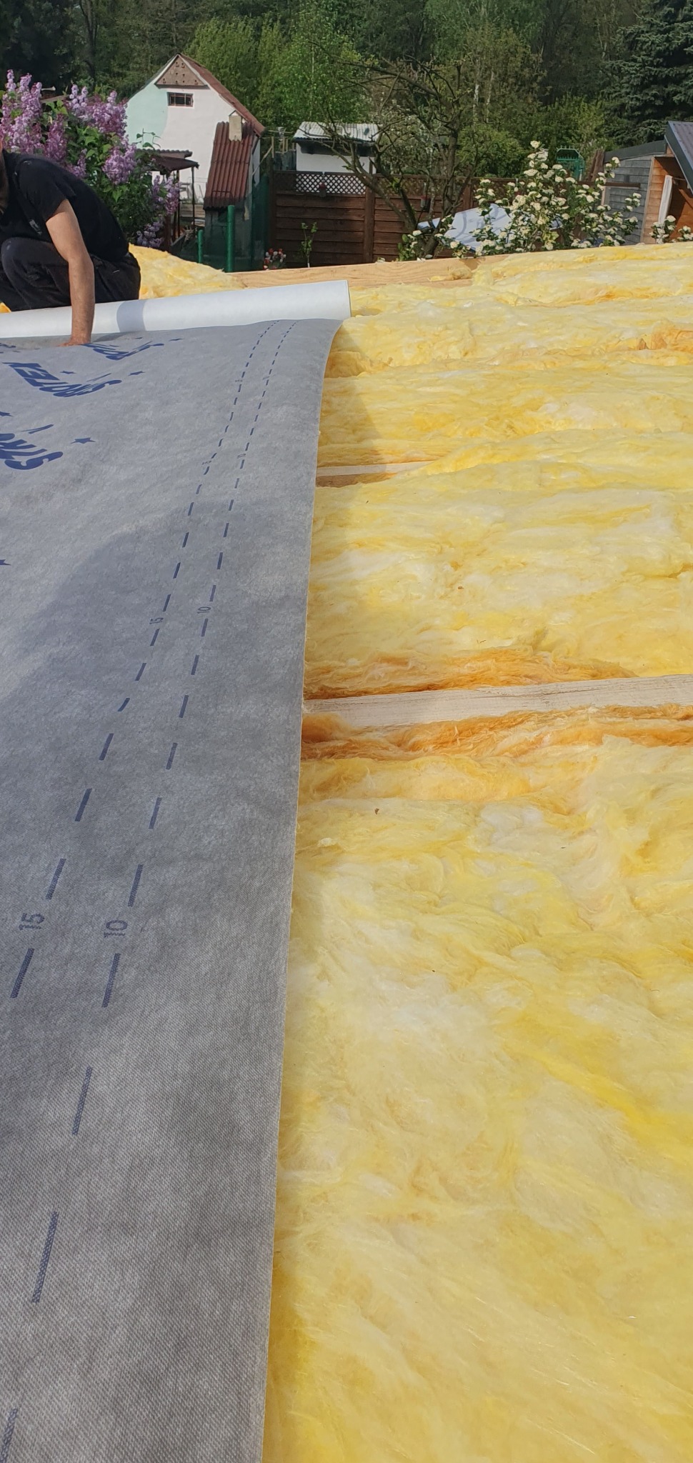 Układanie izolacji z wełny mineralnej i membrany dachowej na drewnianej konstrukcji dachu, widok z góry na częściowo ukończony etap prac.