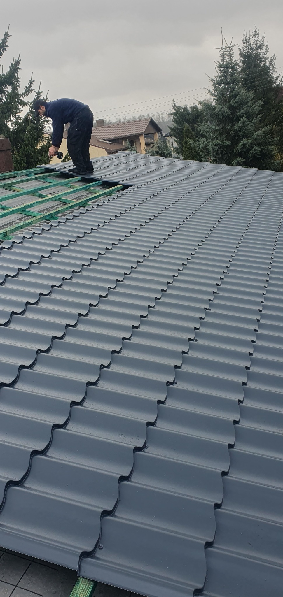 Dekarz montuje szare panele dachowe na konstrukcji drewnianej, widoczny budynek w tle i pochmurne niebo.