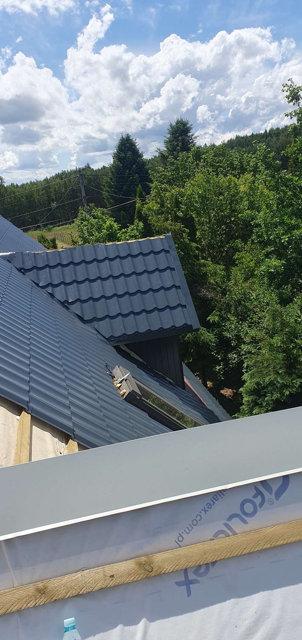 Nowy, grafitowy dach z blachodachówki modułowej, widok z góry na fragment w trakcie montażu okna dachowego, w tle zieleń drzew i błękitne niebo z chmurami.