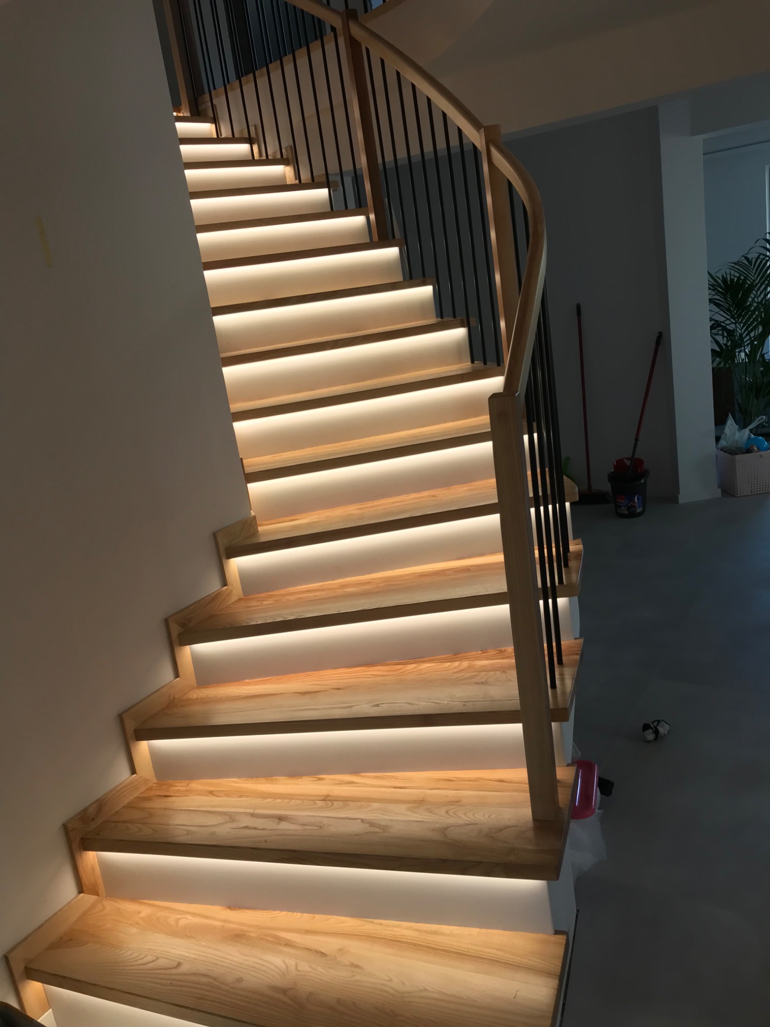 Drewniane schody z podświetleniem LED pod stopniami, czarne metalowe barierki, widok z dołu w górę, jasne drewno, szara podłoga w tle.