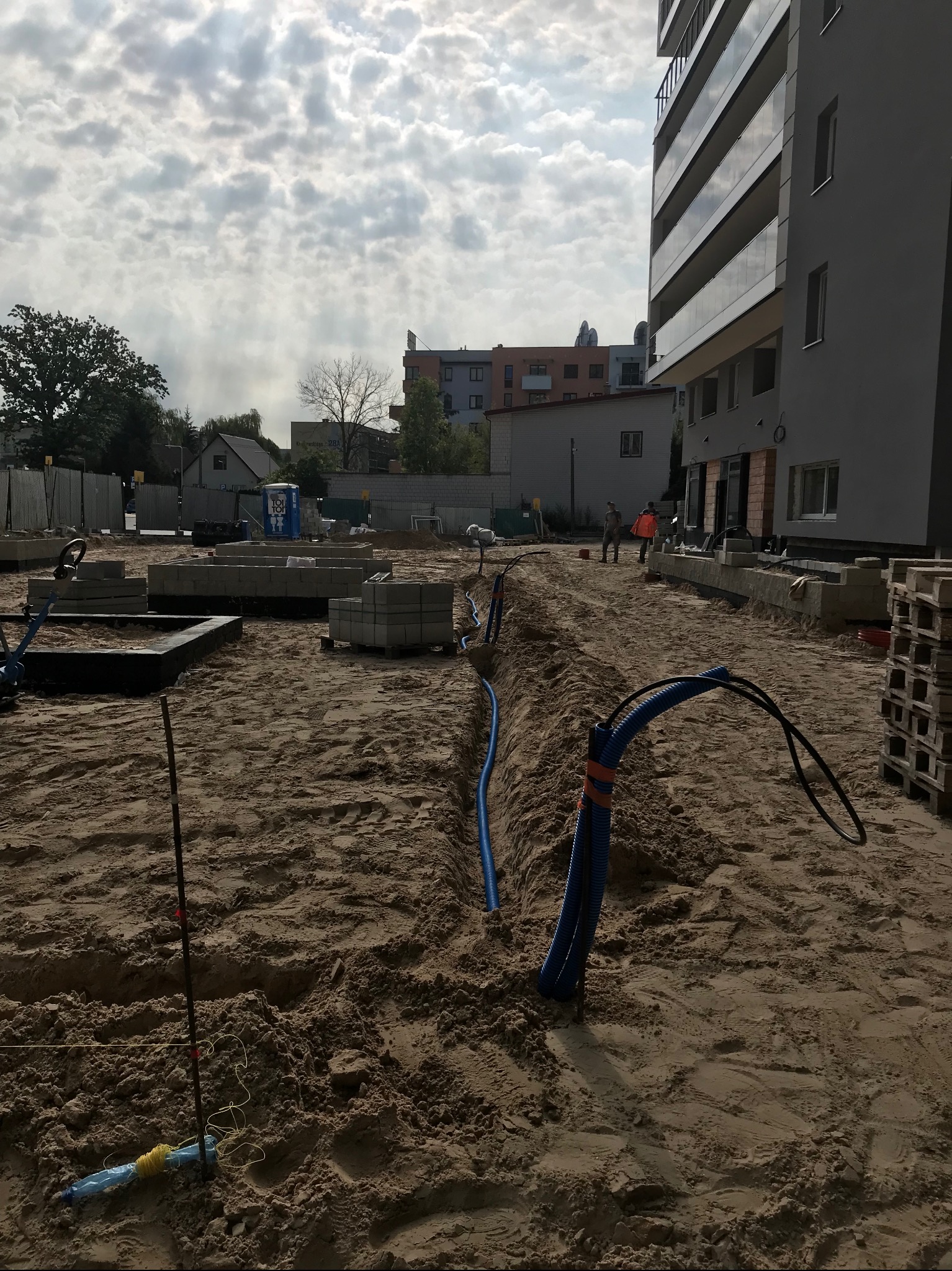 Wkopane w piasek niebieskie rury karbowane z kablami na placu budowy nowego budynku mieszkalnego, widoczni robotnicy w oddali.
