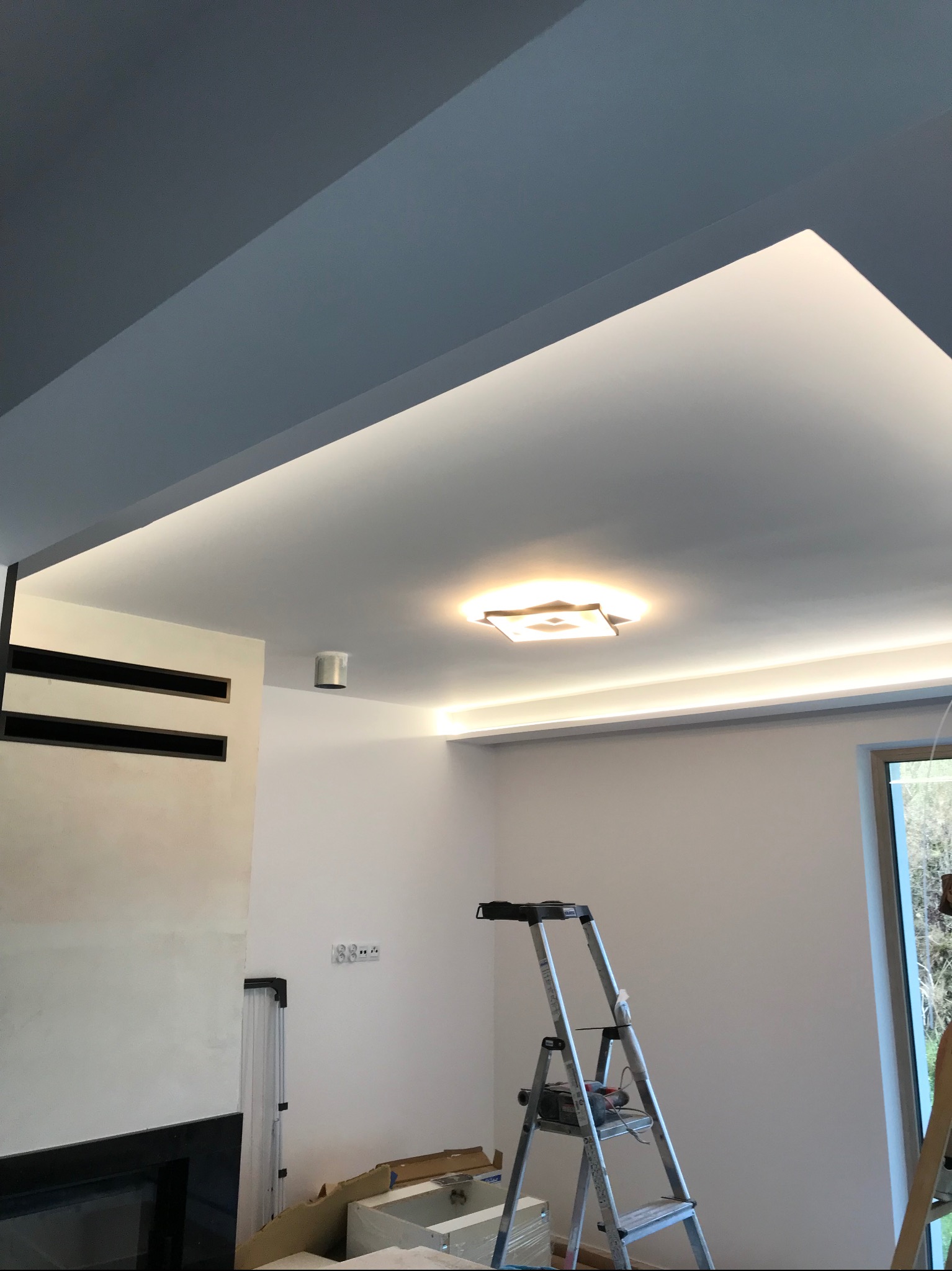 Nowoczesne oświetlenie sufitowe z listwami LED i centralną lampą kwadratową, widoczna drabina malarska i puszka instalacyjna na ścianie, fragment ściany z czarnymi wnękami.