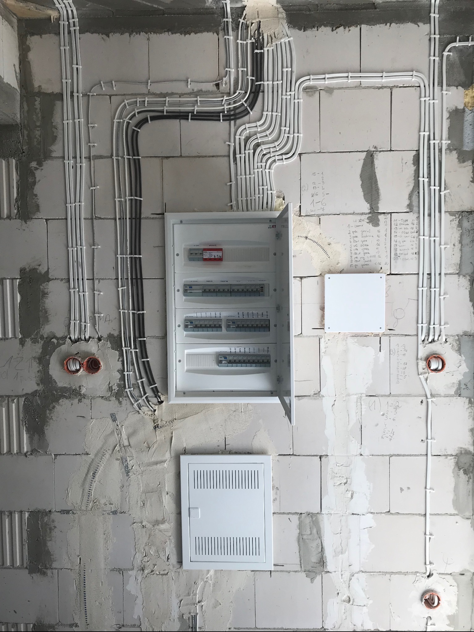 Rozdzielnica elektryczna z otwartymi drzwiami i poprowadzonymi przewodami w białych i czarnych peszlach, zamocowanymi do surowej ściany z betonu komórkowego z widocznymi zapiskami i otworami...
