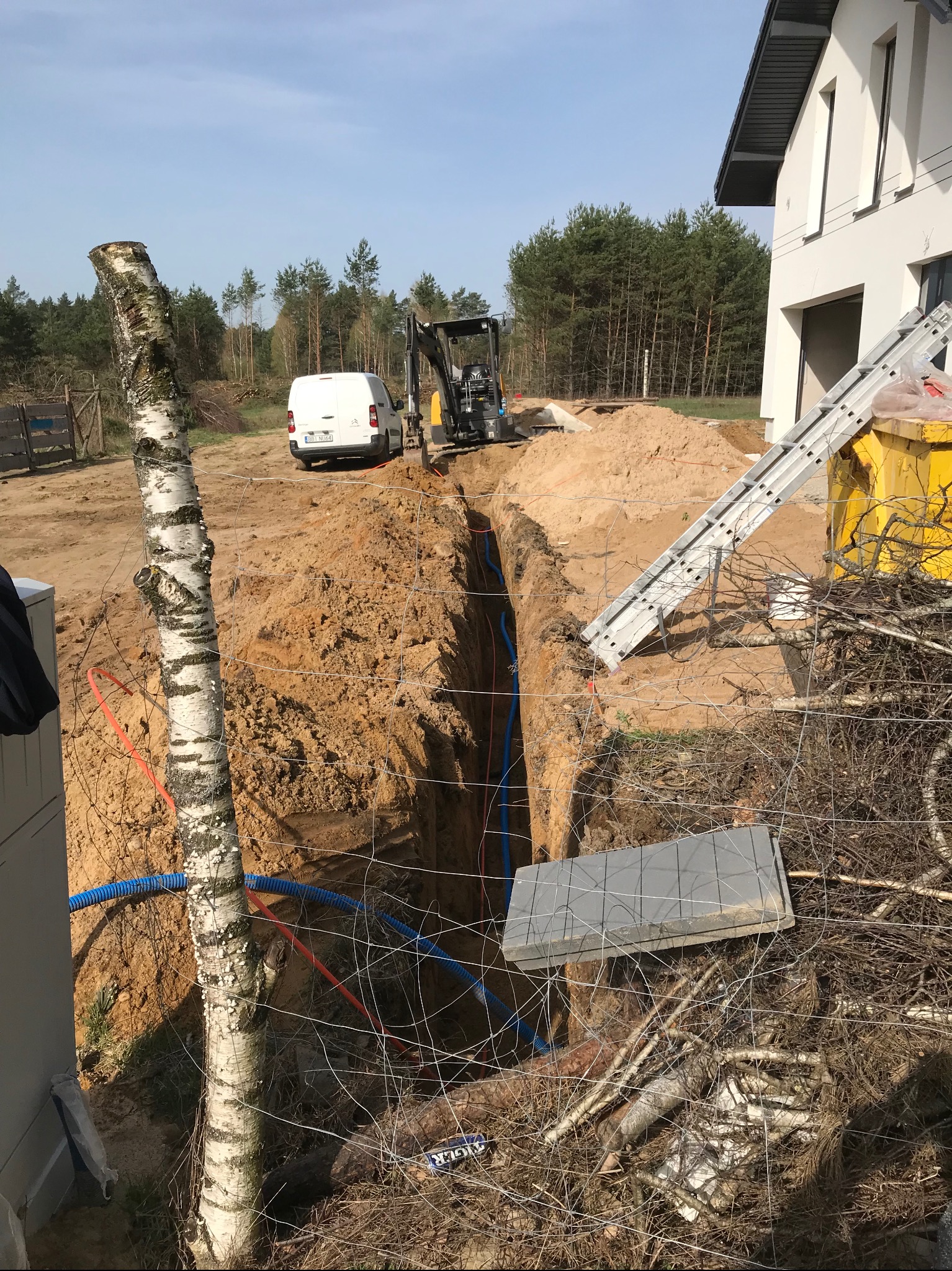 Wykop w ziemi z widocznymi rurami (niebieskie i pomarańczowe) układanymi wzdłuż wykopu, w tle mini koparka i biały samochód dostawczy, obok wykopu leżą płyty chodnikowe i drabina oparta o kontener,...