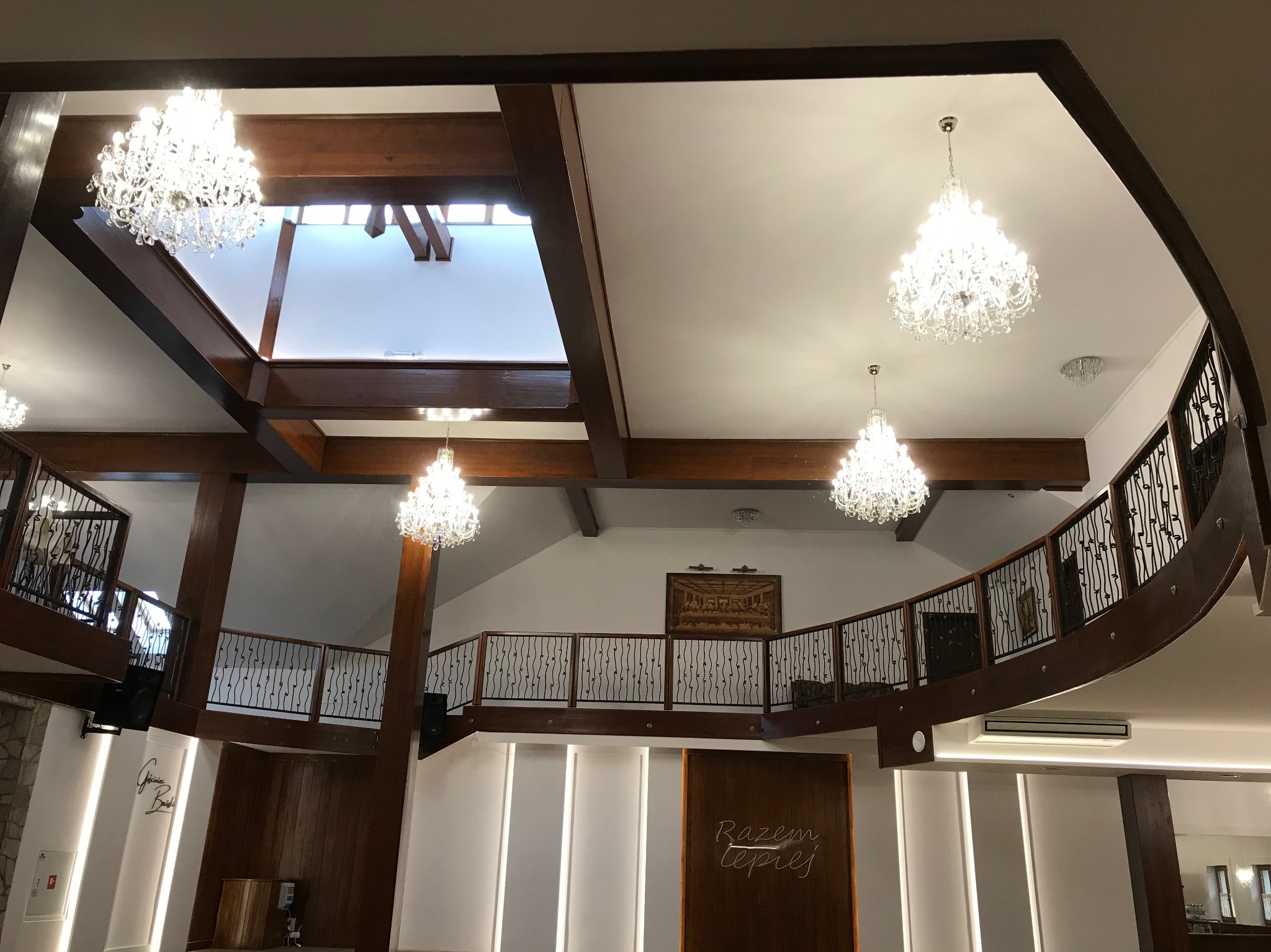 Wnętrze sali bankietowej z drewnianą konstrukcją stropu i balustrad, oświetlone kilkoma kryształowymi żyrandolami, widok z dołu.