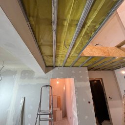 ReNovoPro Oleksandr Shevchenko - Wnętrze w trakcie prac wykończeniowych: sufit z metalową konstrukcją, izolacją z wełny mineralnej i częściowo nałożoną gładzią, ściany po wstępnym szpachlowaniu, drabina aluminiowa i skrzynka...