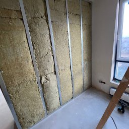 ReNovoPro Oleksandr Shevchenko - Ściana wewnętrzna w trakcie izolacji wełną mineralną, widoczne metalowe profile konstrukcyjne, nieotynkowana podłoga, fragment drabiny i okno w tle.