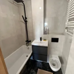 Nowoczesna łazienka po remoncie z czarną armaturą: deszczownica, bateria umywalkowa, szafka pod umywalką w kolorze antracytowym, biała wanna i toaleta, płytki imitujące kamień na ścianach...