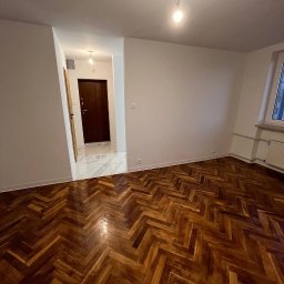 ReNovoPro Oleksandr Shevchenko - Pusty pokój z odnowioną parkietową podłogą ułożoną w jodełkę, świeżo pomalowanymi białymi ścianami i widocznym oknem z grzejnikiem, w tle otwarte drzwi do przedpokoju z marmurową posadzką.