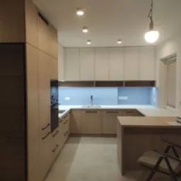 Nowoczesna kuchnia z drewnianymi szafkami dolnymi i białymi górnymi, błękitne płytki nad blatem, wyspa kuchenna z krzesłem, oświetlenie punktowe i wisząca lampa.