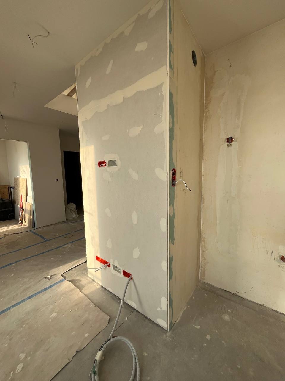Ściana działowa z płyt gipsowo-kartonowych w trakcie szpachlowania, widoczne otwory instalacyjne z wystającymi przewodami elektrycznymi, surowy beton na podłodze z zaznaczonymi liniami, w tle...
