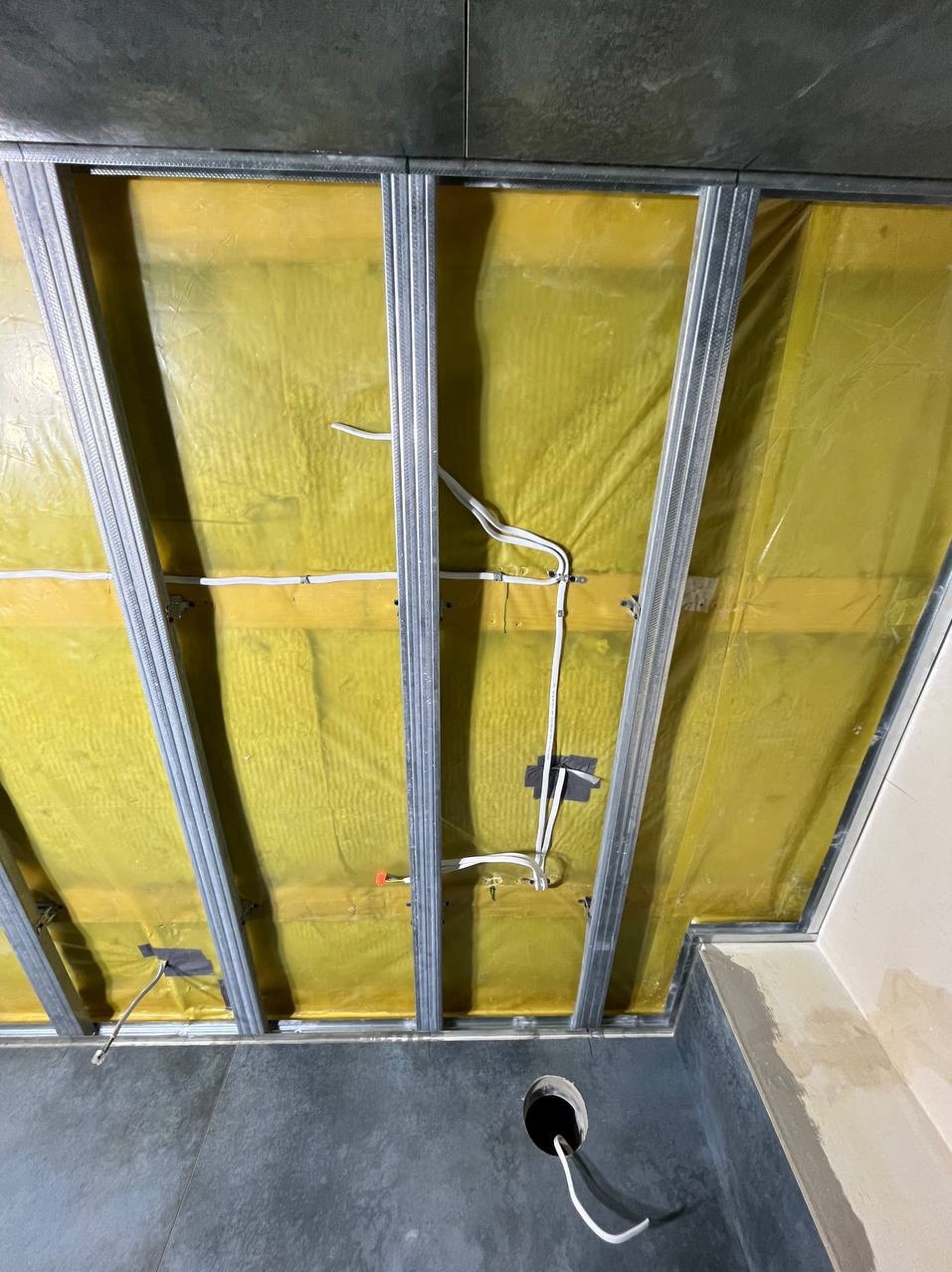 Sufit podwieszany w trakcie montażu: widoczne profile stalowe, żółta wełna mineralna jako izolacja, instalacja elektryczna z przewodami przymocowanymi taśmą, surowy beton na podłodze z otworem...