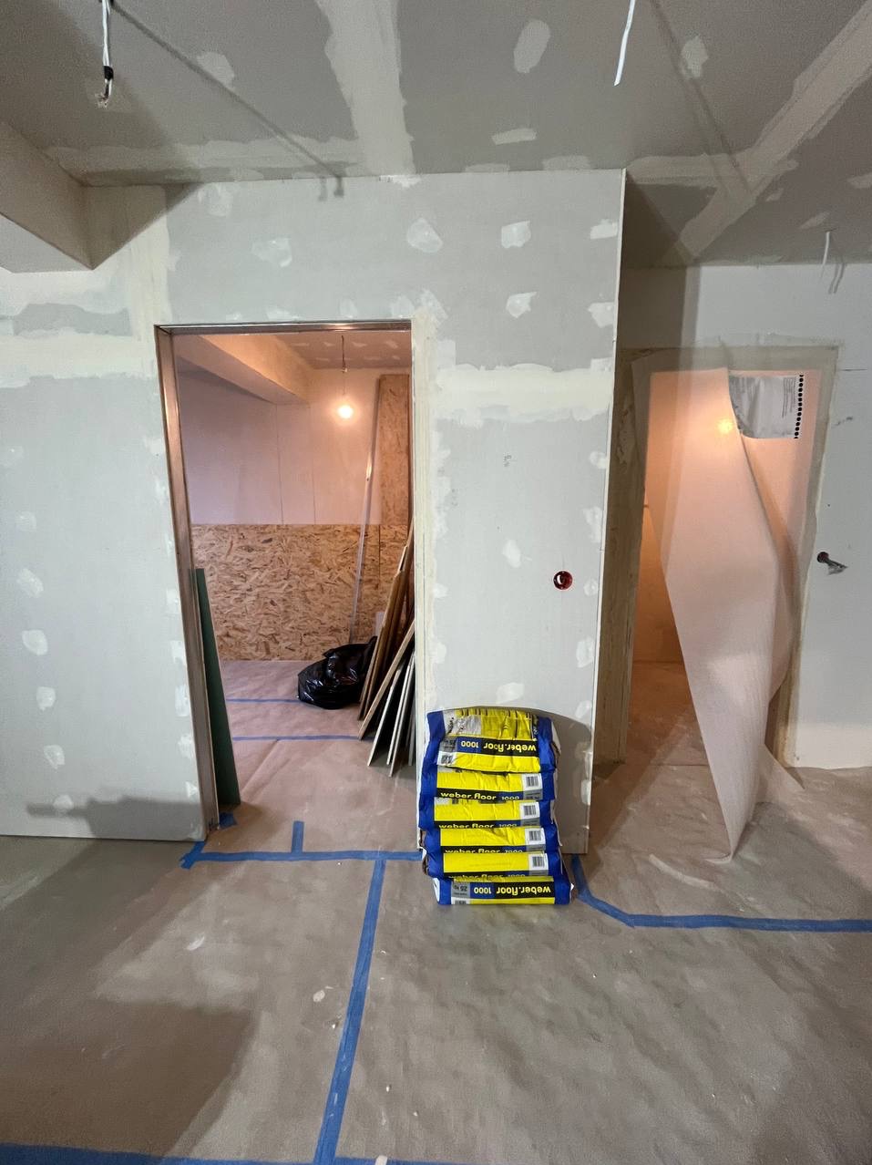 Wnętrze w trakcie prac wykończeniowych: ściany z widocznymi śladami szpachlowania, podłoga zabezpieczona folią i taśmą, stos worków z zaprawą Weber Floor 1000, widoczne materiały budowlane...