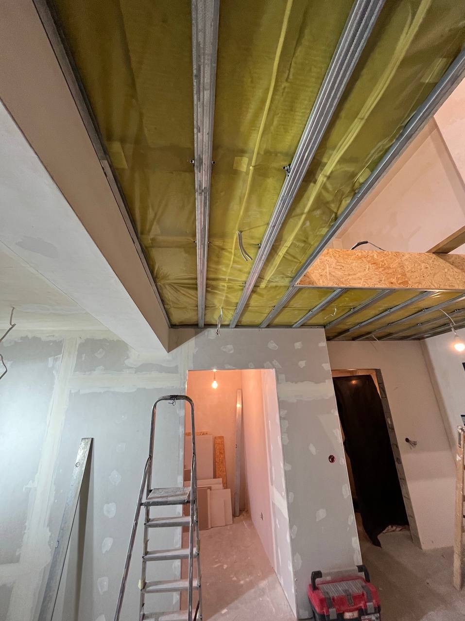 Wnętrze w trakcie prac wykończeniowych: sufit z metalową konstrukcją, izolacją z wełny mineralnej i częściowo nałożoną gładzią, ściany po wstępnym szpachlowaniu, drabina aluminiowa i skrzynka...