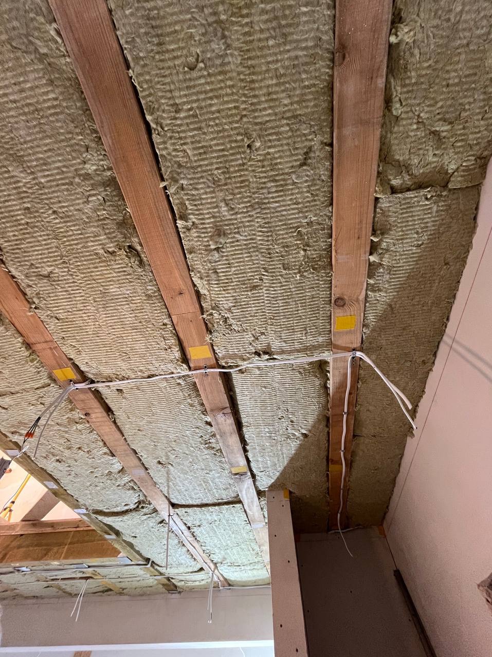 Ocieplenie poddasza wełną mineralną między drewnianymi belkami stropowymi, widoczne przewody elektryczne mocowane do belek żółtą taśmą.