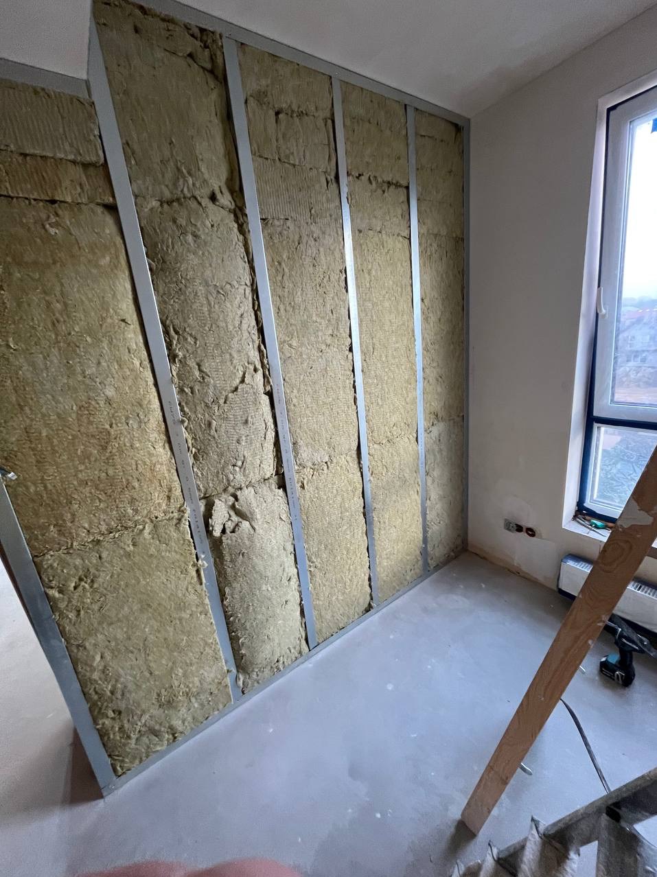 Ściana wewnętrzna w trakcie izolacji wełną mineralną, widoczne metalowe profile konstrukcyjne, nieotynkowana podłoga, fragment drabiny i okno w tle.