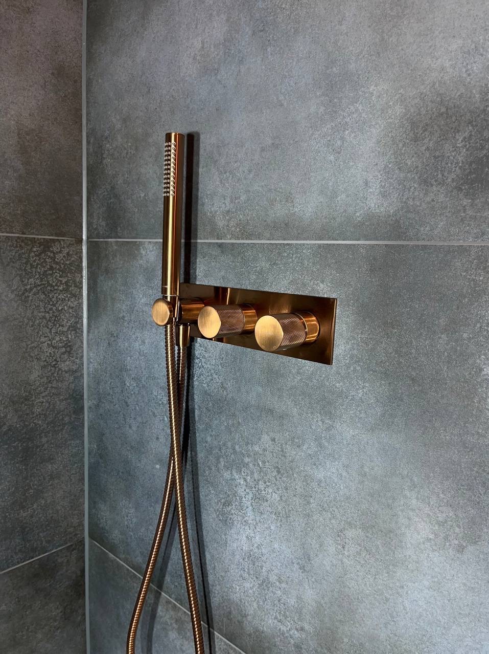 Miedziana bateria prysznicowa podtynkowa z deszczownicą na szarej, teksturowanej ścianie z płytek. Widoczne trzy pokrętła z delikatnym radełkowaniem.