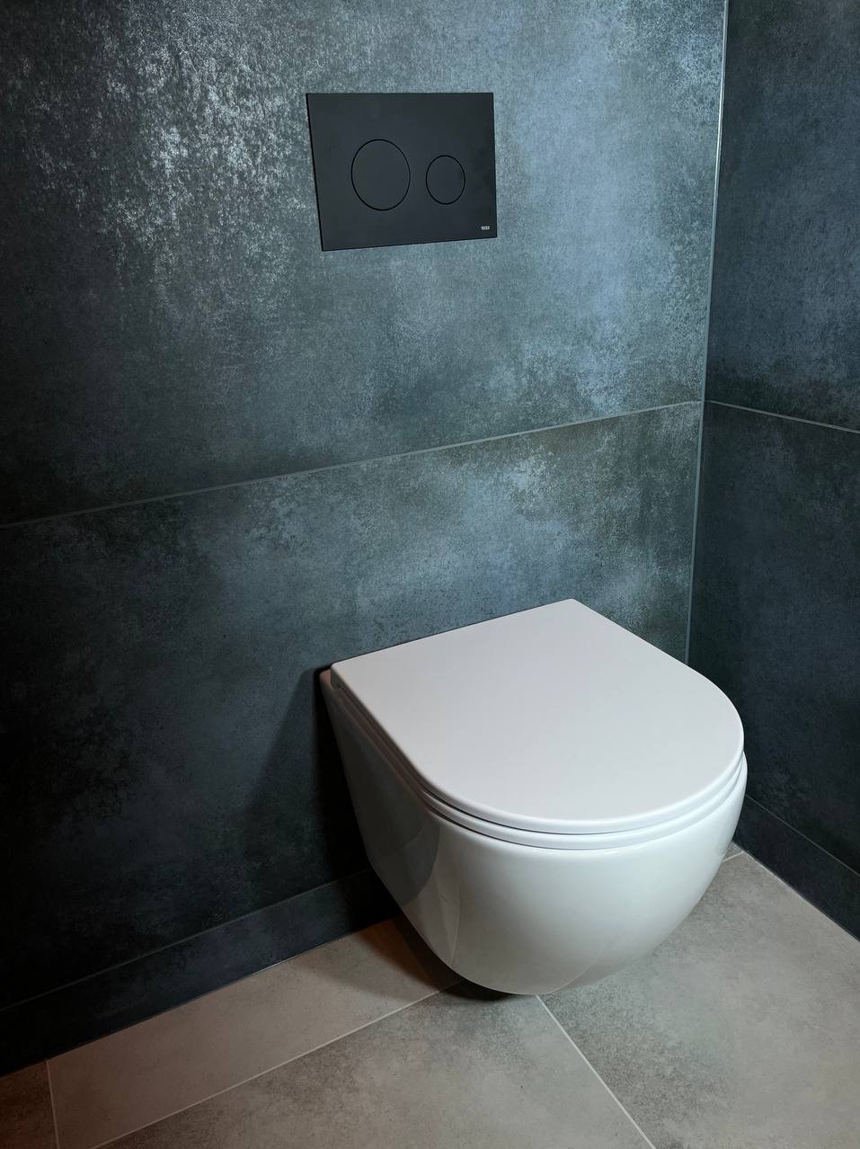 Nowoczesna, podwieszana toaleta z czarnym panelem spłukującym na ścianie z efektem betonu i szarą podłogą z płytek.