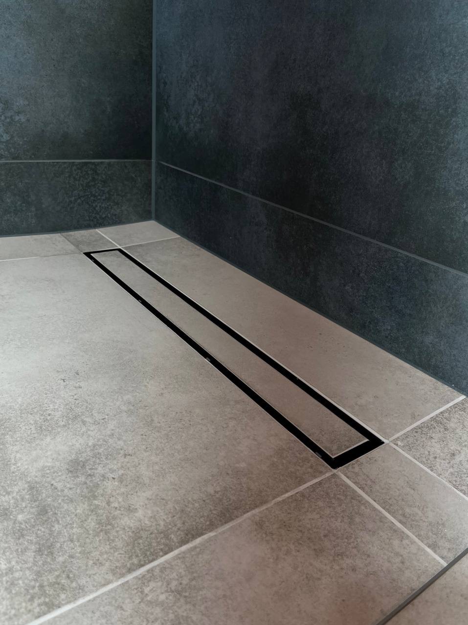 Nowoczesny odpływ liniowy z czarną ramką w jasnoszarej, kwadratowej posadzce łazienki, ciemne płytki na ścianie w tle.