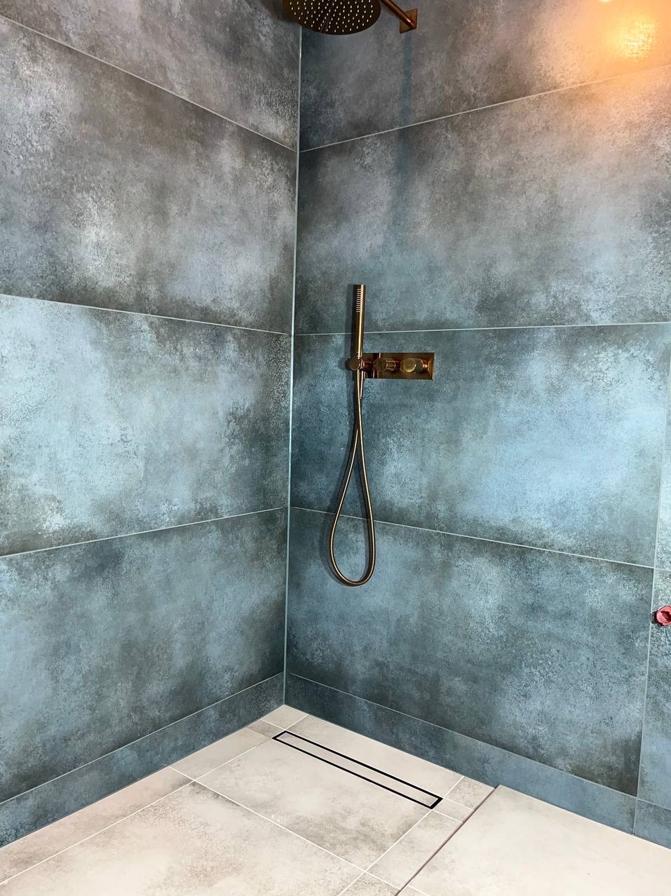 Nowoczesna kabina prysznicowa z szarymi płytkami imitującymi beton, armatura w kolorze miedzianym, odpływ liniowy w podłodze z jasnych płytek.