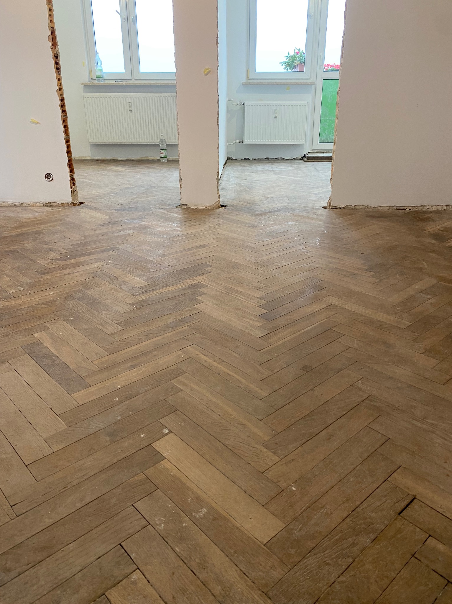 Parkiet ułożony w jodełkę w trakcie remontu, widoczne ślady po pracach wykończeniowych na podłodze i niedokończone ściany z odsłoniętą cegłą, dwa okna z grzejnikami w tle.