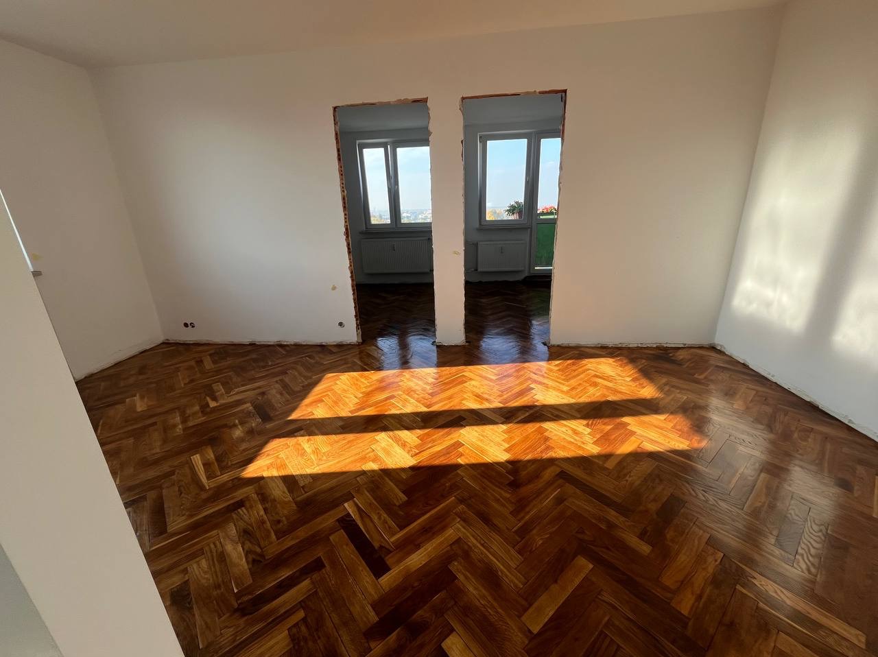 Pokój po remoncie z odnowionym parkietem ułożonym w jodełkę, świeżo pomalowane ściany na biało i przygotowane otwory drzwiowe, wpadające promienie słoneczne.