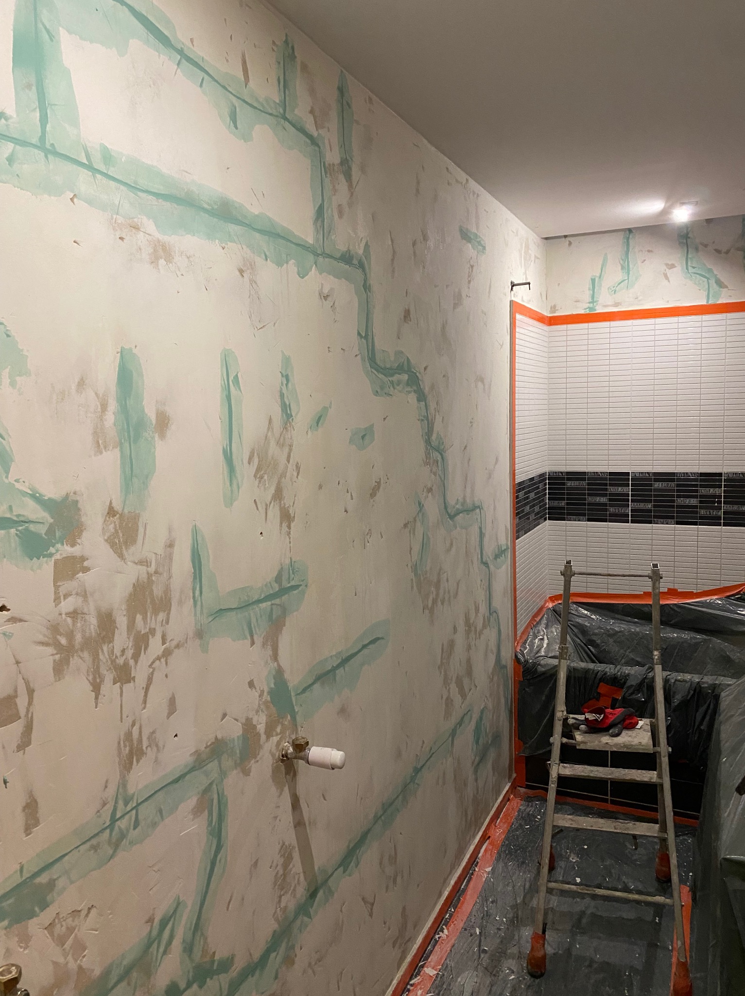 Przygotowanie ścian w łazience do położenia płytek, widoczne pionowe i poziome ślady po taśmie malarskiej zabezpieczającej instalacje, częściowo wykończone płytki na ścianie, drabina w tle...