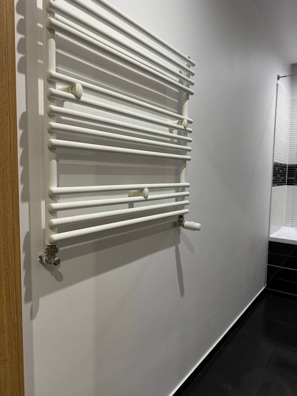 Biały, drabinkowy grzejnik łazienkowy, zamontowany na szarej ścianie, z widocznymi chromowanymi zaworami i fragmentem czarno-białej łazienki w tle.