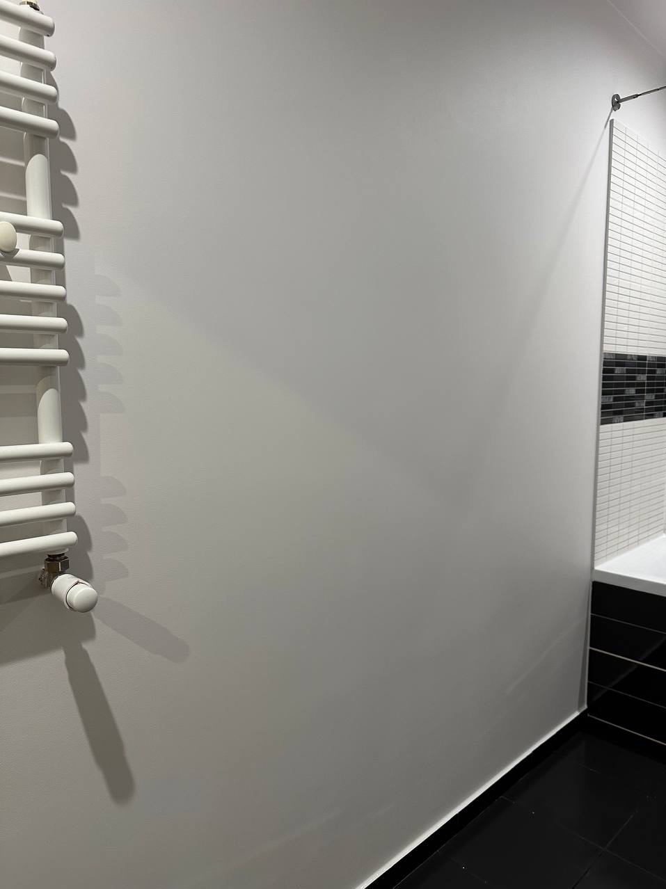 Świeżo pomalowana szara ściana w łazience z białym grzejnikiem drabinkowym po lewej stronie i mozaiką w odcieniach bieli i czerni w prawym górnym rogu, czarna podłoga z płytek.