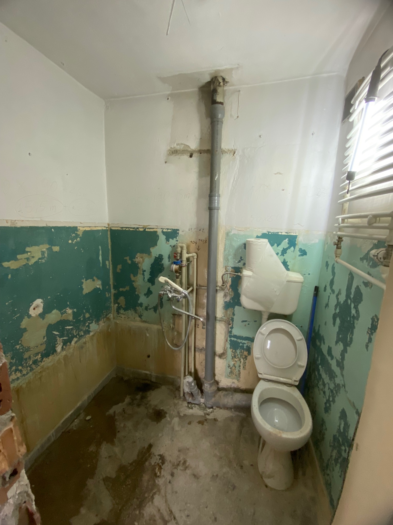 Łazienka w trakcie demontażu, widoczne stare płytki z odpryskami, odsłonięte instalacje hydrauliczne i toaleta z otwartą klapą, ściany z resztkami farby i zapiskami.