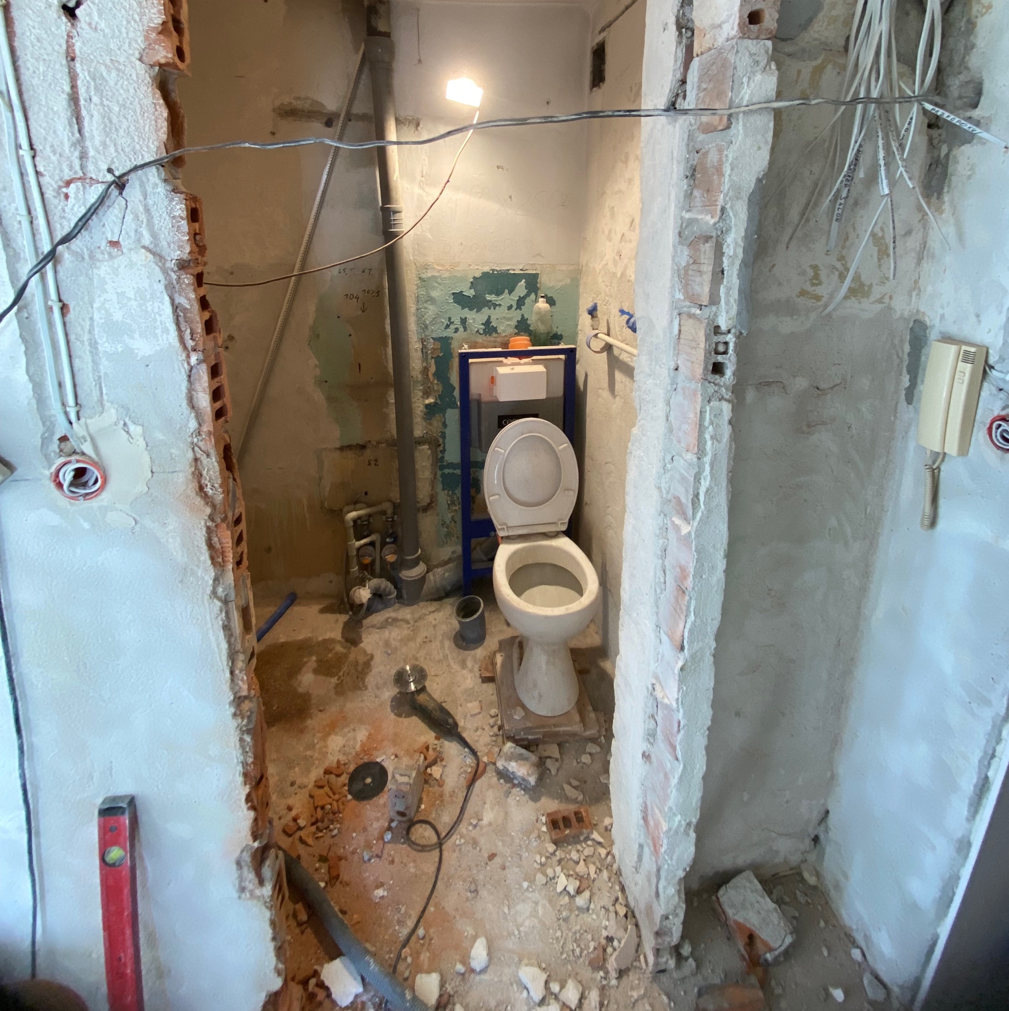 Łazienka w trakcie demontażu: widoczna toaleta, niedokończone ściany z odsłoniętą cegłą, przewody elektryczne, poziomica, szlifierka kątowa i gruz na podłodze.