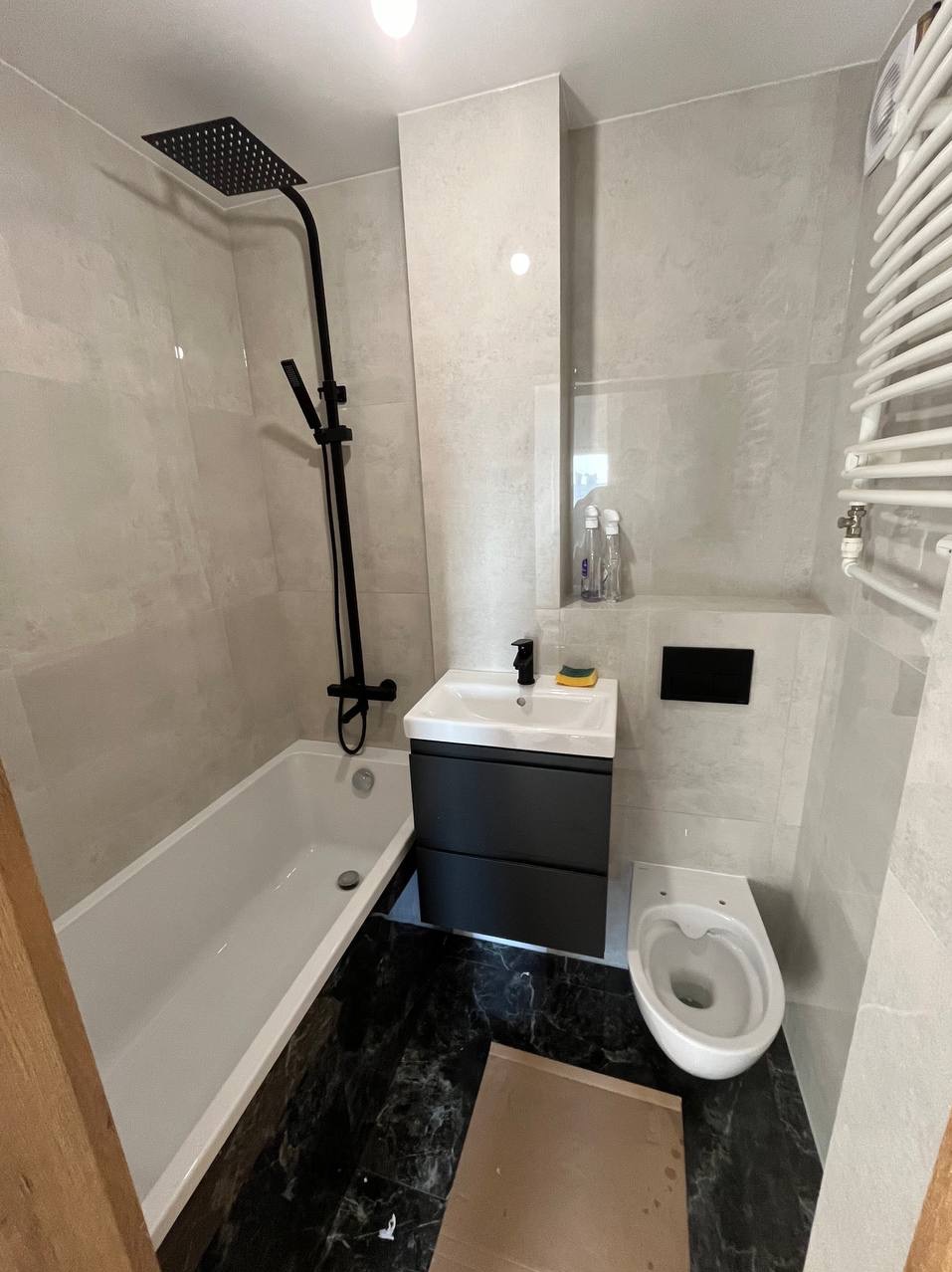 Nowoczesna łazienka po remoncie z czarną armaturą: deszczownica, bateria umywalkowa, szafka pod umywalką w kolorze antracytowym, biała wanna i toaleta, płytki imitujące kamień na ścianach...