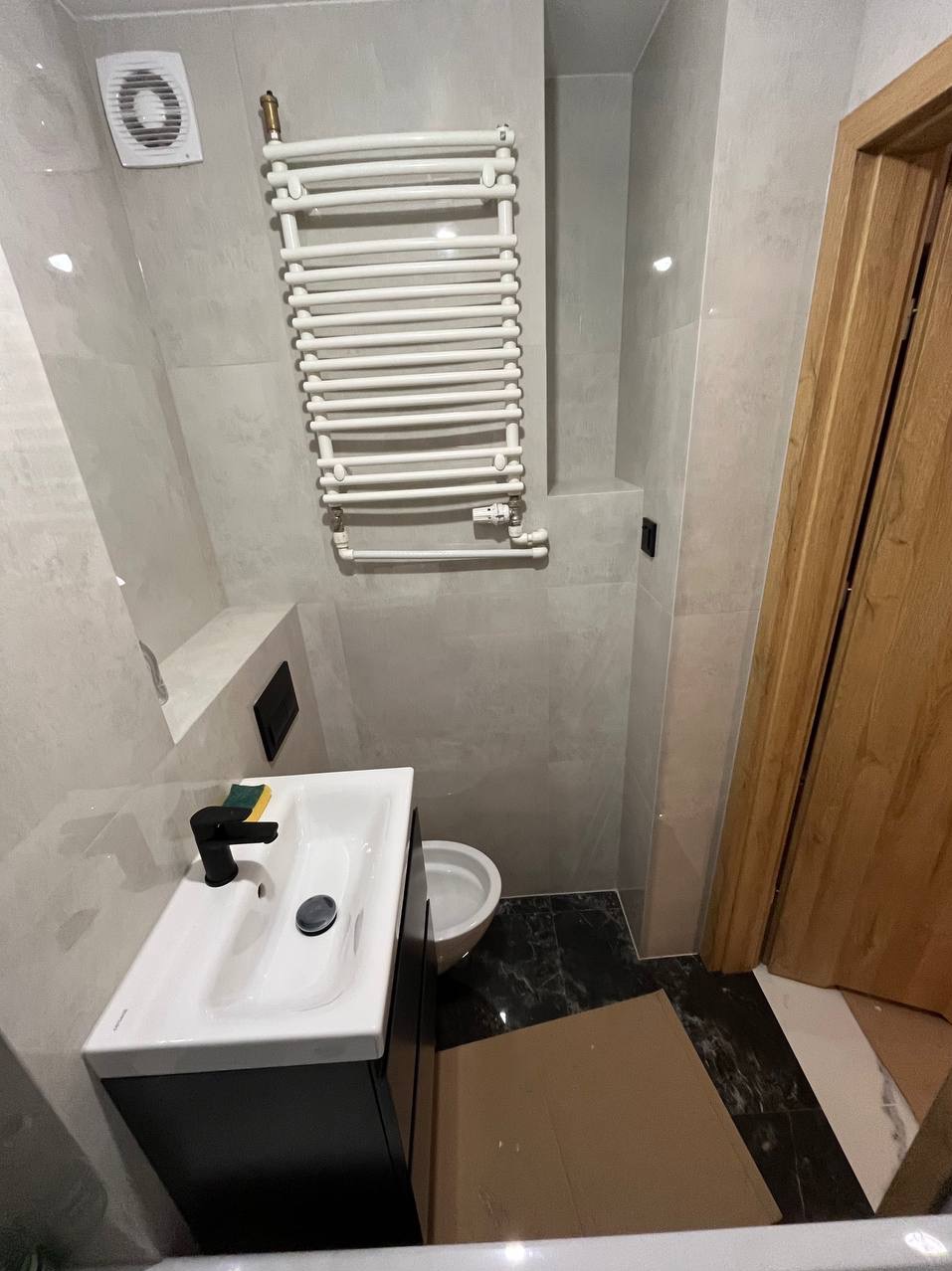 Wykończona łazienka w szarościach z czarnymi akcentami: biała umywalka z czarną armaturą, grzejnik drabinkowy i marmurowe płytki podłogowe, widoczny fragment drewnianych drzwi.