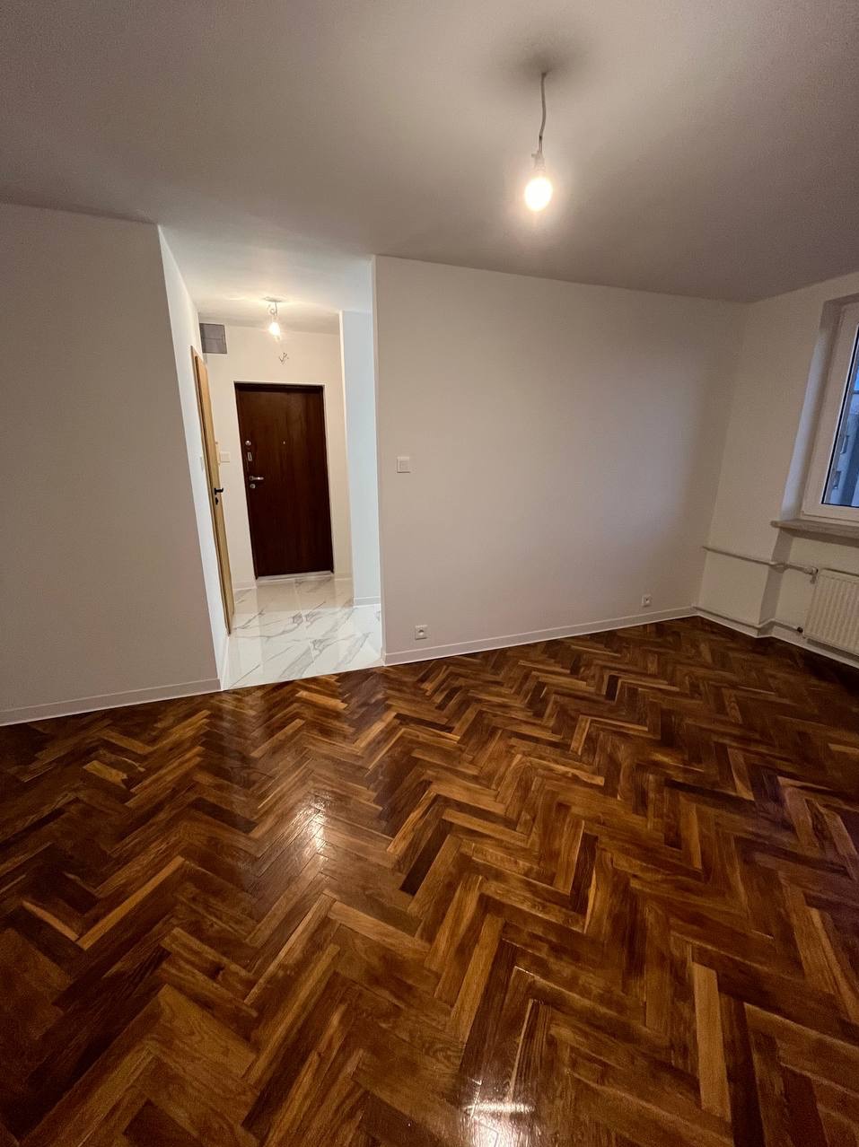 Pusty pokój z odnowioną parkietową podłogą ułożoną w jodełkę, świeżo pomalowanymi białymi ścianami i widocznym oknem z grzejnikiem, w tle otwarte drzwi do przedpokoju z marmurową posadzką.