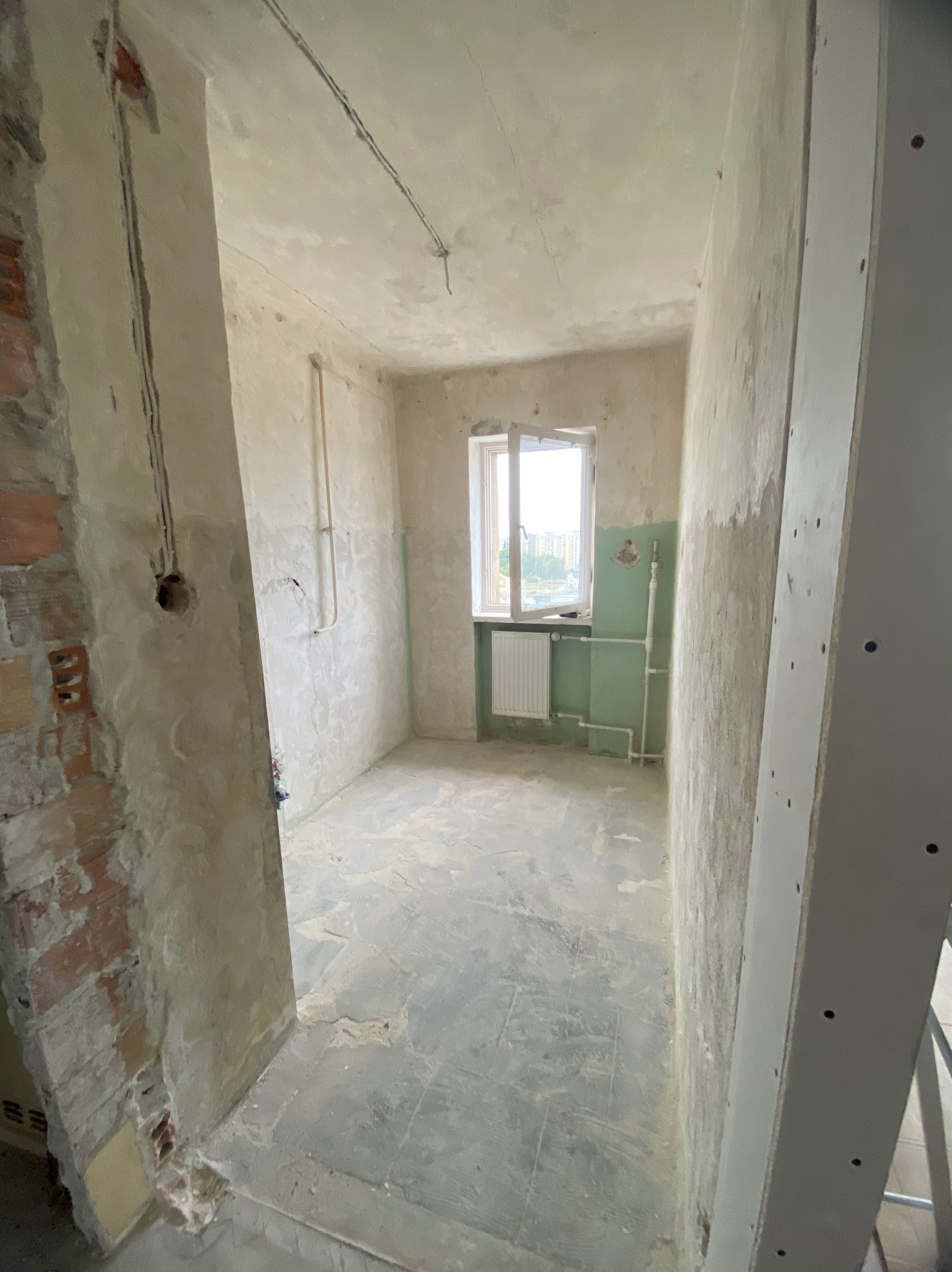 Wnętrze w trakcie prac przygotowawczych do remontu: ściany z resztkami tynku, odsłonięta cegła przy ościeżnicy, grzejnik pod oknem, widoczne instalacje hydrauliczne.