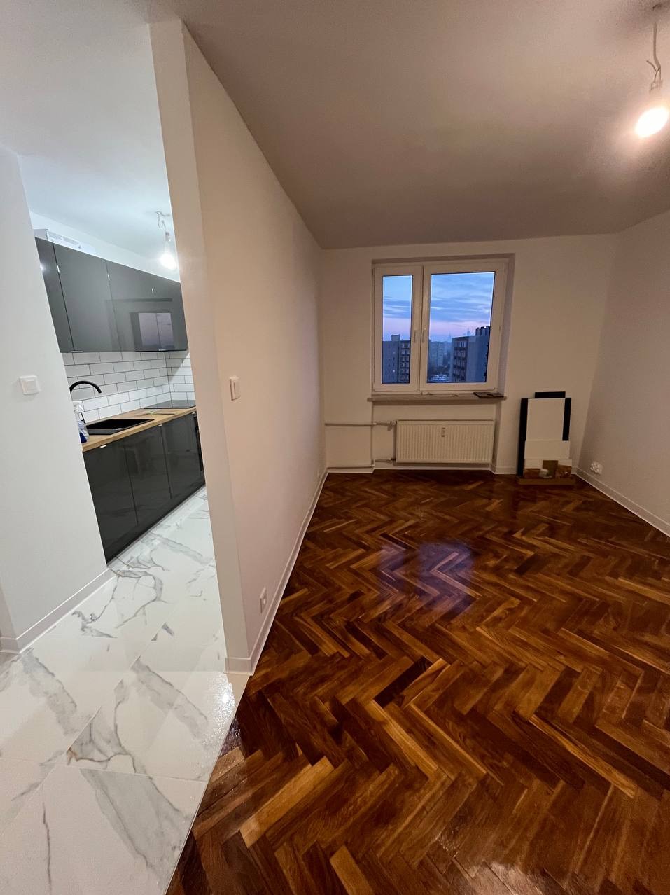 Widok na odnowione mieszkanie: parkiet ułożony w jodełkę, białe ściany i marmurowa podłoga w otwartej kuchni z szarymi szafkami.