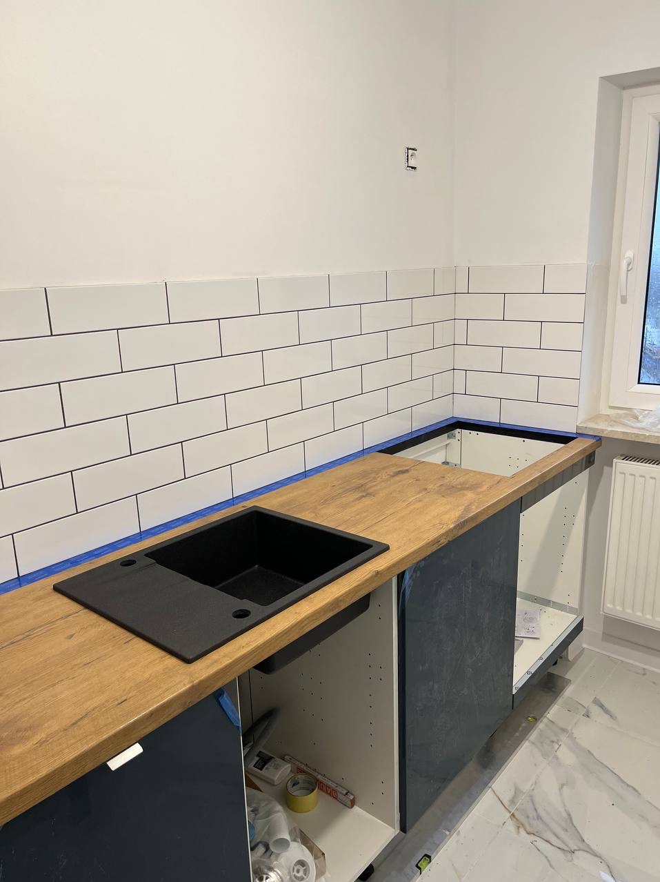 Montaż czarnego zlewozmywaka w drewnianym blacie kuchennym, widoczne płytki na ścianie zabezpieczone niebieską taśmą malarską, szafki kuchenne w kolorze granatowym, realizacja w Ząbkach.
