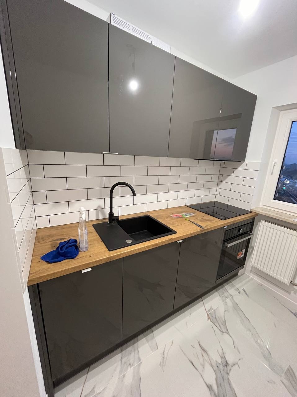 Nowoczesna kuchnia z szarymi, lakierowanymi szafkami, drewnianym blatem, czarnym zlewem i baterią, białymi płytkami na ścianie oraz marmurową podłogą, widoczna butelka ze spryskiwaczem i niebieska...