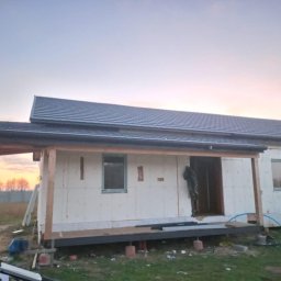 Vitalii Povtoreyko - Wznoszenie domu modułowego z widoczną izolacją ścian zewnętrznych i fragmentem dachu pokrytego szarą dachówką, na tle zachodzącego słońca.