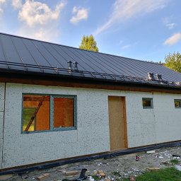 Vitalii Povtoreyko - Budynek w trakcie budowy, ocieplony styropianem, z zamontowanymi oknami i drzwiami, pokryty blachą trapezową w kolorze grafitowym, widoczne rynny i kominki wentylacyjne.